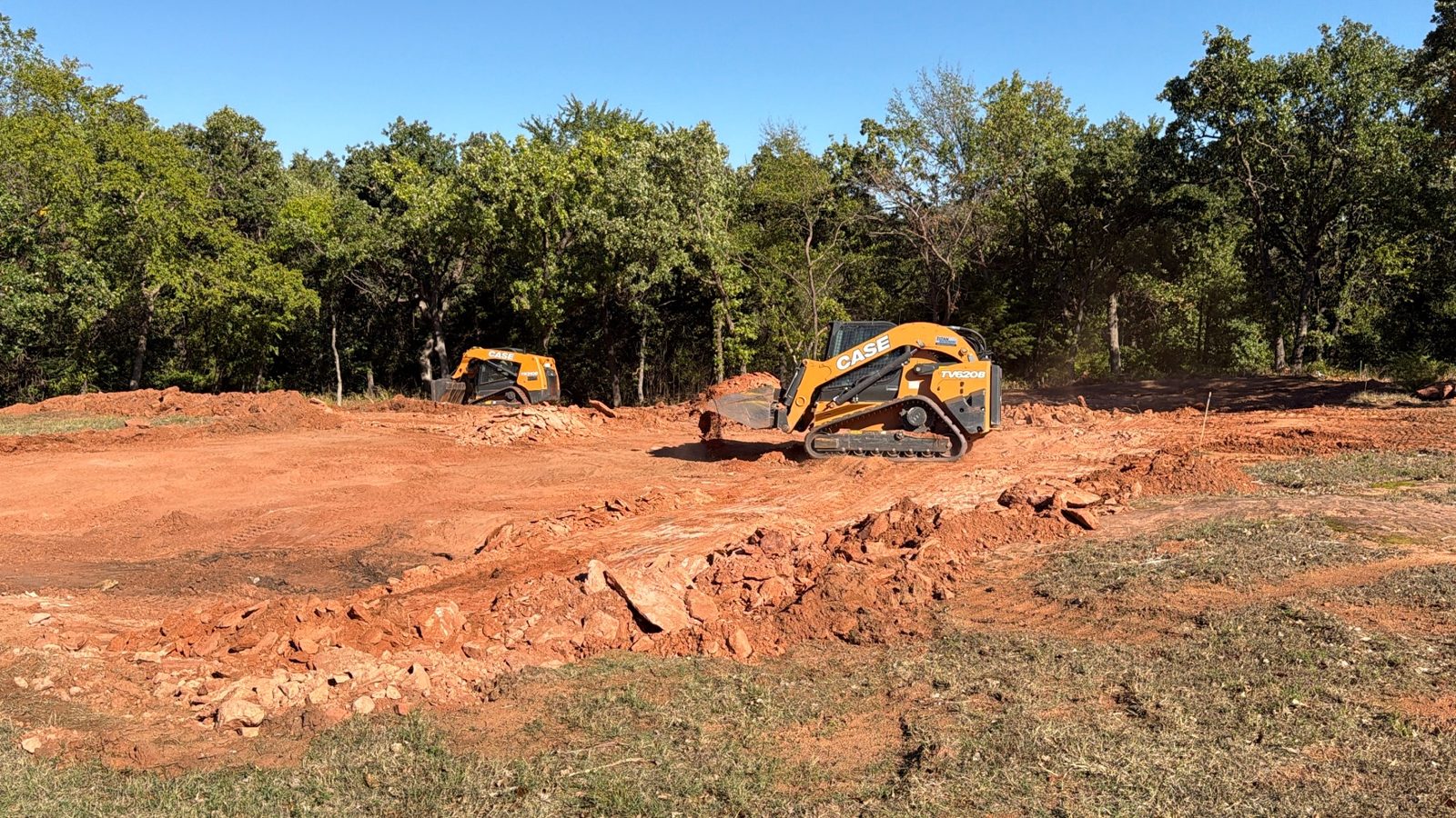 Track loaders clearing rock and red dirt on a wooded lot in Logan County